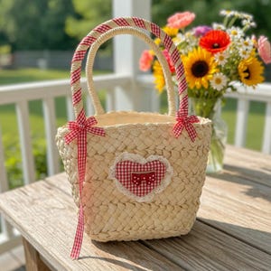 May include: A woven straw basket with a heart-shaped patch. The basket has a handle wrapped in red and white gingham ribbon, with matching bows. A red and white gingham heart is centered on the front, trimmed with white lace.
