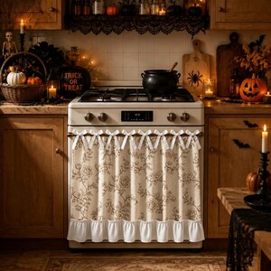Puede incluir: Una cortina de horno beige con un patrón floral marrón y un borde inferior con volantes blancos. La cortina está sujeta al mango del horno con una serie de lazos de tela blanca, cubriendo el frente del electrodoméstico en una cocina.