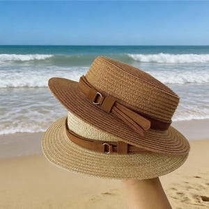 May include: Two straw hats with brown leather bands and metal buckles. The hats are tan and brown, with a wide brim and a flat top. The hats are held up against a beach background with the ocean in the distance.