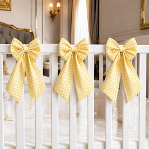 May include: Three decorative bows made of yellow and white gingham fabric attached to the vertical slats of a white wooden crib. Each bow features a central knot and two long, hanging tails that drape downward along the crib railing.