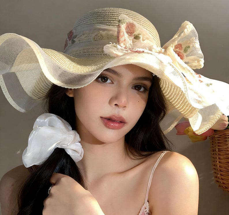 K&ouml;nnte beinhalten: Ein breitkrempiger Strohhut mit floralem Design und einer gro&szlig;en Schleife. Der Hut hat einen cremefarbenen Mesh-&Uuml;berzug und ist mit rosa und gr&uuml;nen floralen Akzenten verziert. Ein stilvolles Accessoire zum Sonnenschutz.