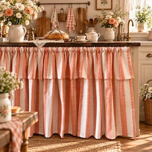 May include: A kitchen sink skirt featuring a vertical striped pattern in coral orange and white. The fabric is gathered at the top with a ruffled valance overlay, creating a layered appearance that covers the lower cabinet area of the kitchen island.