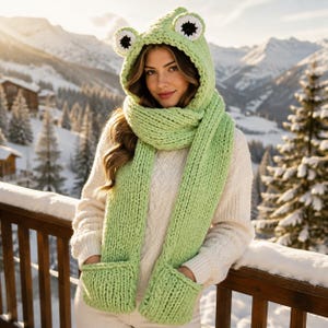 May include: A light green chunky knit hooded scarf featuring two frog eyes on top of the hood and two integrated pockets at the ends of the scarf. The accessory is crafted with a thick ribbed texture and is designed to cover the head and neck.