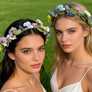 May include: Two women wearing handmade crochet flower crowns. The crowns feature a variety of colorful flowers in shades of blue, yellow, pink, and purple, with green leaves. The women are in front of a green grassy background.
