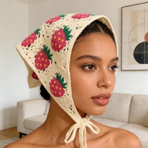 May include: A cream-coloured crochet headscarf featuring a repeating pattern of pink strawberries with green leaves. The headscarf is tied under the chin with a cream-coloured string. The strawberries have yellow seeds.