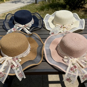 K&ouml;nnte beinhalten: Vier Sonnenh&uuml;te aus Stroh in Marineblau, Creme, Beige und Rosa, jeweils mit breiter, gewellter Krempe und dekorativer Schleife. Die H&uuml;te haben ein Band mit floralen Akzenten und eine transparente Mesh-Borte.