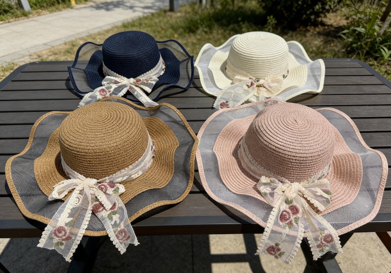 K&ouml;nnte beinhalten: Vier Sonnenh&uuml;te aus Stroh in Marineblau, Creme, Beige und Rosa, jeweils mit breiter, gewellter Krempe und dekorativer Schleife. Die H&uuml;te haben ein Band mit floralen Akzenten und eine transparente Mesh-Borte.