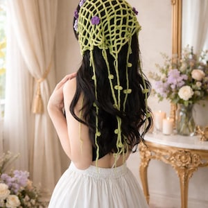 May include: A green crocheted head covering with purple floral accents. The headpiece has long, dangling strands with small green embellishments. The person is wearing a black top and a white skirt. The background includes a gold mirror and a table with flowers.