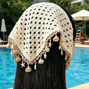 May include: Cream-colored crochet headscarf with a square shape and a decorative border. The border is adorned with small seashells and starfish charms. The headscarf is worn over dark hair, with a swimming pool in the background.