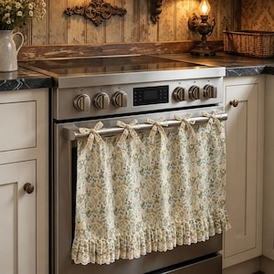 May include: A cream-colored fabric oven door cover featuring a delicate botanical print of wildflowers, greenery, and small blue butterflies. The cover is attached to the oven handle with five fabric ties and features a ruffled bottom edge.