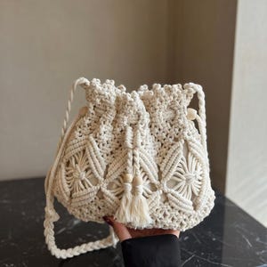Può includere: Borsa in macramè color crema con chiusura a coulisse e nappa. La borsa presenta un design intrecciato con motivi floreali e dettagli di perline di legno. La borsa ha una tracolla.