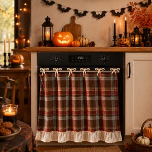 May include: A brown, red, and blue plaid oven curtain featuring a ruffled cream-coloured hem. The fabric is gathered and attached to the oven handle with six small, tied cream-coloured fabric bows, creating a decorative covering for the appliance.