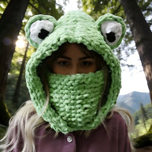 May include: A light green, handmade frog hood with large, white and black eye details. The hood covers the head and neck, with a knitted scarf-like section covering the lower face. The soft, textured knitwear is a whimsical accessory.