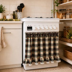 May include: A white kitchen stove featuring a plaid fabric cover draped over the oven door. The fabric has a red, green, and cream tartan pattern, accented with small bows along the top edge and a white lace trim along the bottom hem.