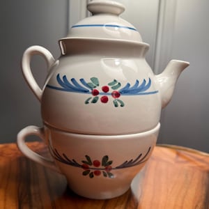 May include: A white ceramic teapot and teacup set. The teapot has a lid and spout, and the teacup is stacked beneath it. Both pieces are decorated with blue and green floral designs and red berries. The set is on a wooden surface.