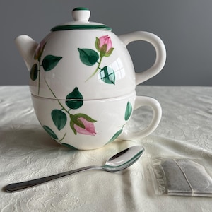 May include: A white ceramic teapot and teacup set with a pink rose and green leaf design. A silver spoon and a tea bag are next to the set. The teapot has a green rim and lid.