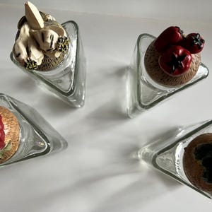 May include: Four clear glass triangular bottles with decorative stoppers. The stoppers feature various designs, including a bunny, red berries, and a dark green leafy design. The bottles are arranged on a white surface.