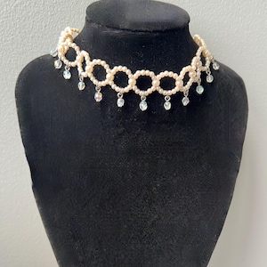 May include: A cream-colored pearl choker necklace with a circular design. The necklace features small pearls and dangling iridescent crystal accents. The necklace is displayed on a black mannequin.