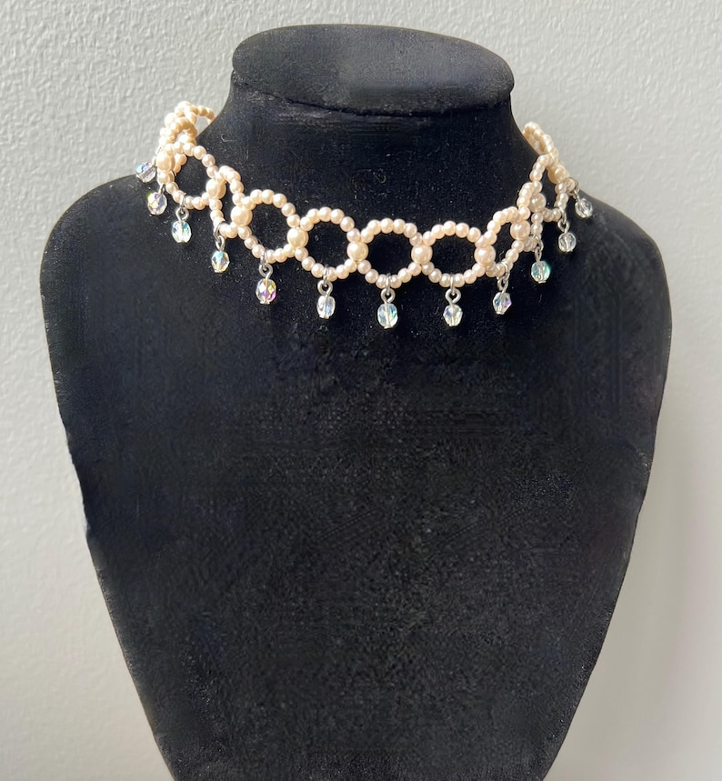 May include: A cream-colored pearl choker necklace with a circular design. The necklace features small pearls and dangling iridescent crystal accents. The necklace is displayed on a black mannequin.