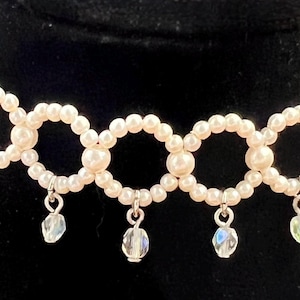 May include: A cream-colored pearl choker necklace with a series of connected circles. Each circle is adorned with small pearls. Clear, iridescent teardrop-shaped crystals dangle from the bottom of the necklace.