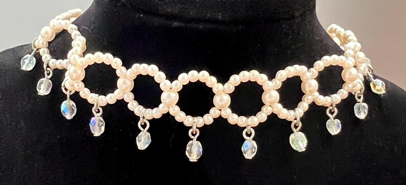 May include: A cream-colored pearl choker necklace with a series of connected circles. Each circle is adorned with small pearls. Clear, iridescent teardrop-shaped crystals dangle from the bottom of the necklace.
