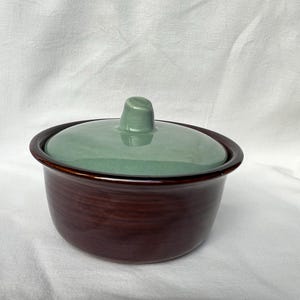 May include: A ceramic serving bowl with a lid. The bowl is a deep brown color with a glossy finish. The lid is a light green color with a small handle. The bowl is sitting on a white surface.