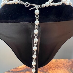 May include: A multi-strand pearl necklace with a silver clasp and a dangling pearl chain. The necklace is displayed on a black velvet bust, showcasing its elegant design and classic appeal. The pearls are white and round.
