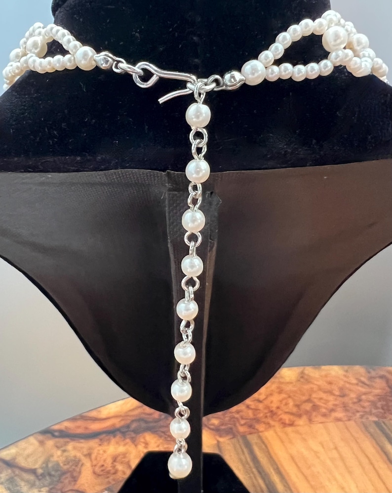 May include: A multi-strand pearl necklace with a silver clasp and a dangling pearl chain. The necklace is displayed on a black velvet bust, showcasing its elegant design and classic appeal. The pearls are white and round.