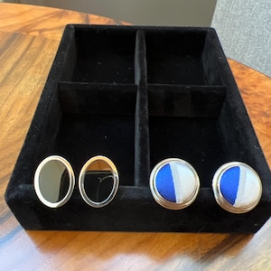 May include: A black velvet jewelry tray with four compartments holds two pairs of cufflinks. One pair features black oval insets with gold-colored frames. The other pair has blue and white striped fabric insets with gold-colored frames.