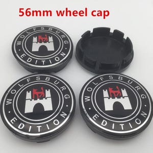 Puede incluir: Cuatro tapacubos circulares de 56 mm con acabado negro y borde plateado. Cada tapa presenta el texto Wolfsburg Edition y un emblema central que representa un castillo blanco con la silueta de un lobo rojo. Una tapa se muestra del revés.