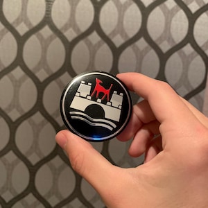 Puede incluir: Un emblema redondo en blanco y negro con un diseño de perro rojo sobre un puente blanco. El emblema se sostiene en una mano sobre un fondo gris estampado. El emblema tiene un acabado brillante.