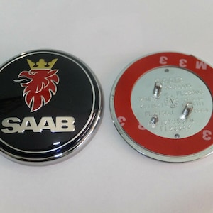 May include: Two round Saab car emblems. One is black with a silver border, featuring a red griffin and the word "SAAB" in silver. The other emblem's back is silver with a red border.