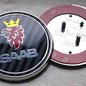 May include: Two-piece car emblem with a carbon fiber pattern. The emblem features the SAAB logo in silver, a red griffin with a yellow crown, and a silver border. The second piece has mounting studs and a red border.