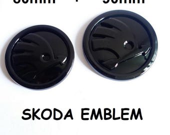 May include: Two glossy black Skoda emblem badges, one 80mm and one 90mm in diameter, with a stylized bird logo. The text "SKODA EMBLEM" is printed below the badges. The badges are circular and have a raised design.