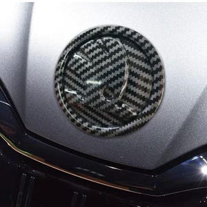 Könnte beinhalten: Ein kreisförmiges Autoemblem mit Carbonfaserstruktur. Das Emblem hat ein schwarz-graues Muster und befindet sich auf einer silbernen Motorhaube. Dies ist ein Autozubehör.