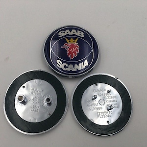 Könnte beinhalten: Drei Automobil-Embleme. Das obere Emblem ist rund mit blauem Hintergrund, dem Wort "SAAB" über einem roten Greif und dem Wort "SCANIA" darunter. Zwei schwarz umrandete Embleme befinden sich darunter, mit dem Wort "SAAB" und Zahlen.