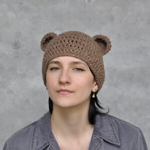 May include: A brown crocheted beanie with bear ears. The beanie is made with a textured stitch and has a simple design.