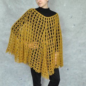 A mustard yellow crochet poncho with a loose, open weave. The poncho has a fringe trim and is worn over a black turtleneck.