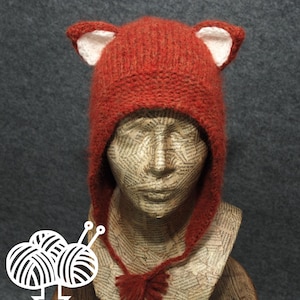 May include: A handmade, rust-colored knit hat with fox ears. The hat features white inner ear details and ear flaps that extend down to a tassel. The hat is displayed on a mannequin head with a newspaper collage design.