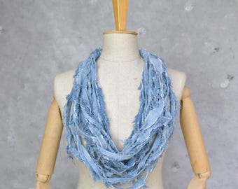 May include: A light blue infinity scarf made of shredded fabric, displayed on a mannequin. The scarf has a frayed, textured appearance, with visible white threads and a soft, casual style. The scarf is a circular design.