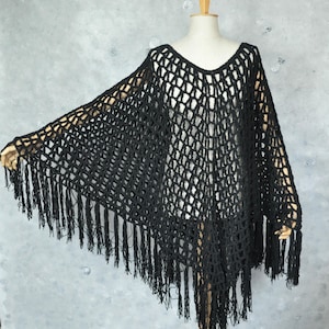 Puede incluir: Un poncho negro de crochet con cuello en V y largas borlas. El poncho se exhibe en un maniquí, mostrando su diseño drapeado y su textura. Las borlas cuelgan a lo largo del borde inferior.
