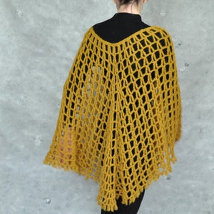 Peut inclure: Un poncho en crochet jaune moutarde avec un motif en dentelle. Le poncho est port&eacute; par une personne aux cheveux violets et un col roul&eacute; noir.