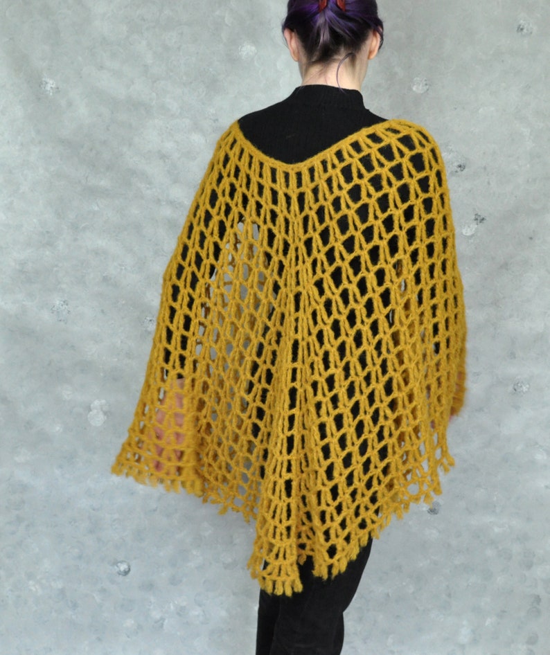 Peut inclure: Un poncho en crochet jaune moutarde avec un motif en dentelle. Le poncho est port&eacute; par une personne aux cheveux violets et un col roul&eacute; noir.