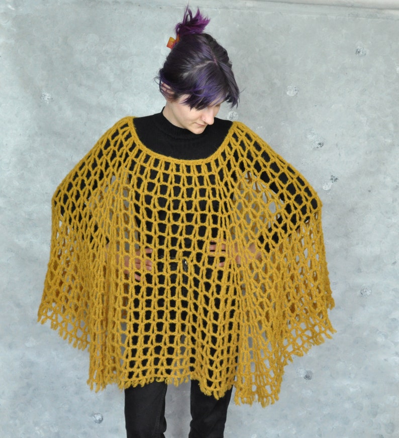 Peut inclure: Un poncho en crochet jaune moutarde &agrave; la maille l&acirc;che et ouverte. Le poncho a une bordure en franges et est port&eacute; par une personne aux cheveux violets.