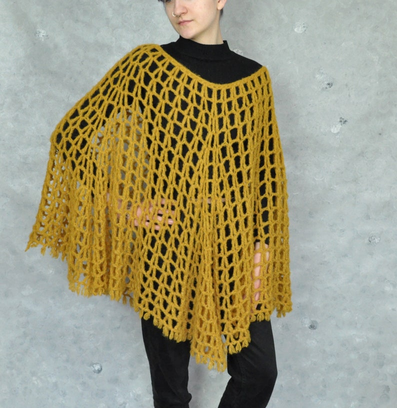 A mustard yellow crochet poncho with a loose, open weave. The poncho has a fringe trim and is worn over a black turtleneck.