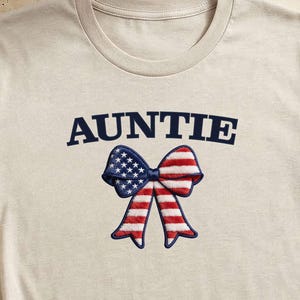 Puede incluir: Una camiseta beige de manga corta con la palabra AUNTIE en letras mayúsculas azul oscuro sobre un gráfico de un lazo con el patrón de la bandera estadounidense en rojo, blanco y azul. La camiseta tiene cuello redondo y un dobladillo anudado.