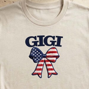 Könnte beinhalten: Ein beiges kurzärmeliges T-Shirt mit dem Schriftzug GIGI in dunkelblauen Buchstaben über einer Schleifengrafik. Die Schleife ist mit den roten, weißen und blauen Sternen und Streifen der amerikanischen Flagge gemustert. Das T-Shirt ist unten geknotet.