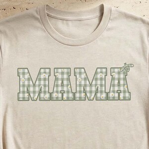May include: A beige short-sleeve t-shirt featuring the word MAMA printed in a block font across the chest. The letters have a sage green and white gingham check pattern with small white flower accents throughout the design.