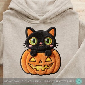 May include: A light beige hooded sweatshirt featuring an embroidered design of a black cat with large green eyes sitting inside an orange jack-o'-lantern pumpkin. The cat has pink cheeks and black whiskers, while the pumpkin has a yellow carved face.