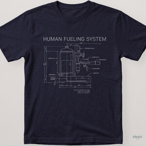 May include: Navy blue t-shirt with a white schematic design labeled "HUMAN FUELING SYSTEM." The diagram resembles a machine with various components and measurements. The shirt has a crew neck and short sleeves.